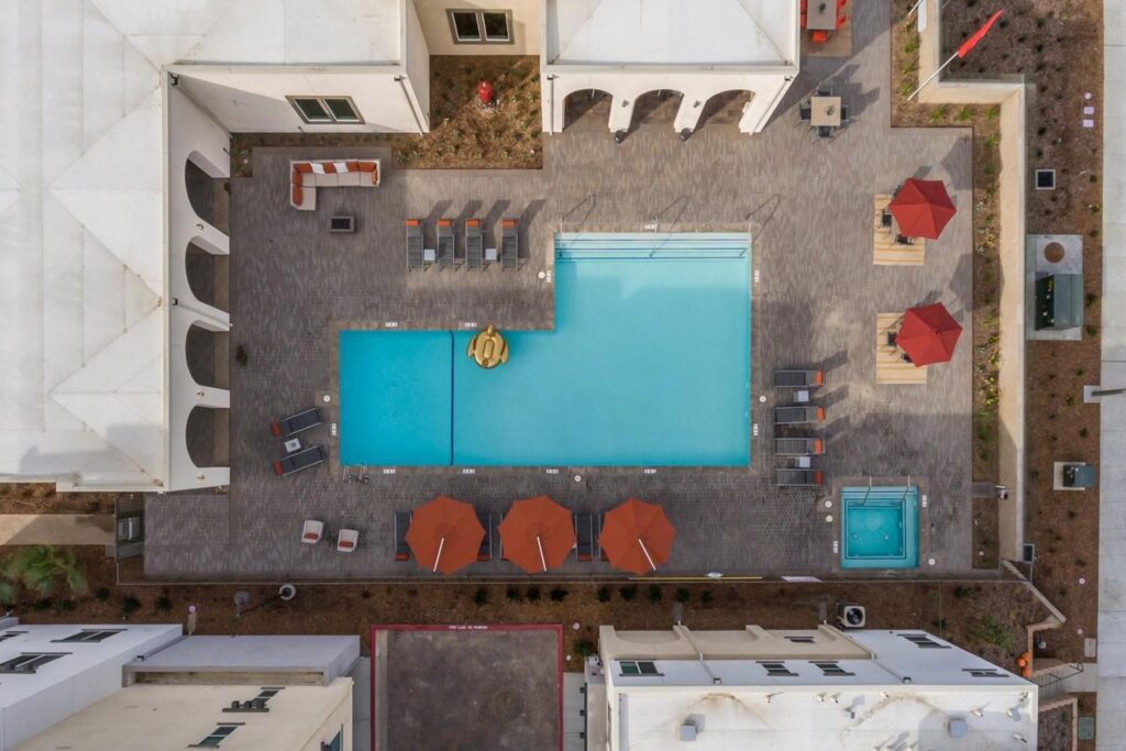 drone shot of the pool with multiple sun loungers, large umbrellas with table sets underneath them and a sofa set.