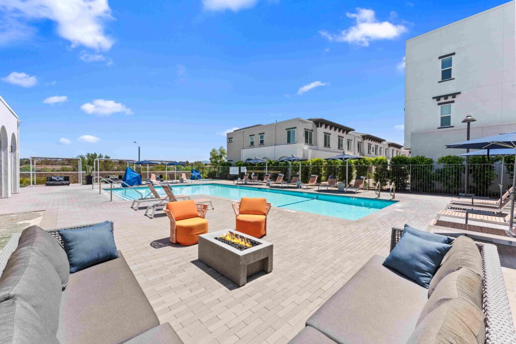 Outdoor pool area with lounge chairs, blue umbrellas, and a fire pit surrounded by cushioned seating.