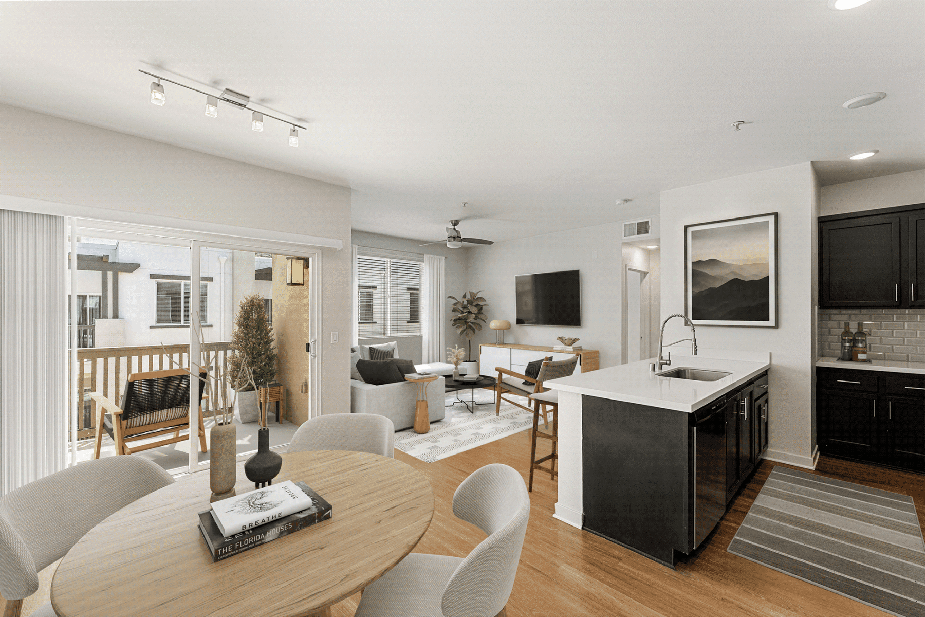 Open-concept apartment with a dining area in the foreground, a living room with a couch and television in the center, and a kitchen with dark cabinetry and a white island to the right.