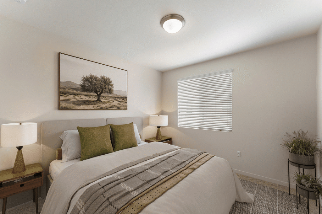 A neutral-toned bedroom with a bed featuring olive green pillows and a patterned throw, flanked by two lamps on nightstands, with framed wall art of a tree and a window with blinds.