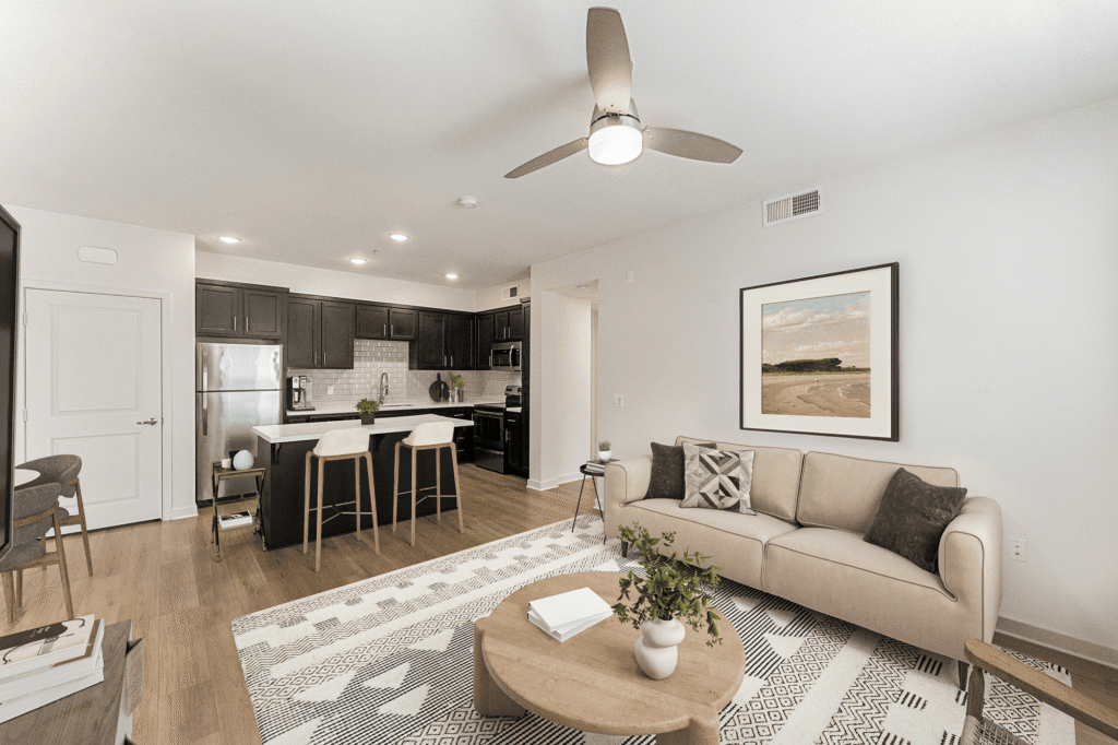 A modern open-concept apartment with a kitchen featuring dark cabinets and a breakfast bar, alongside a living area with a beige sofa, a round wooden coffee table, and a framed beach landscape print on the wall.