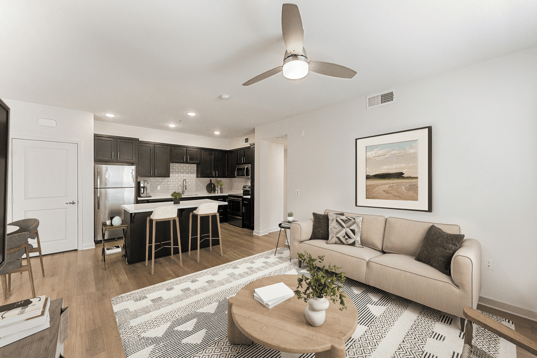 A modern open-concept apartment with a kitchen featuring dark cabinets and a breakfast bar, alongside a living area with a beige sofa, a round wooden coffee table, and a framed beach landscape print on the wall.