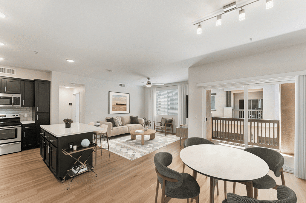 Open-concept kitchen, living room, and dining area featuring a kitchen island, beige sofa, round coffee table, and a dining table with four chairs overlooking a balcony.