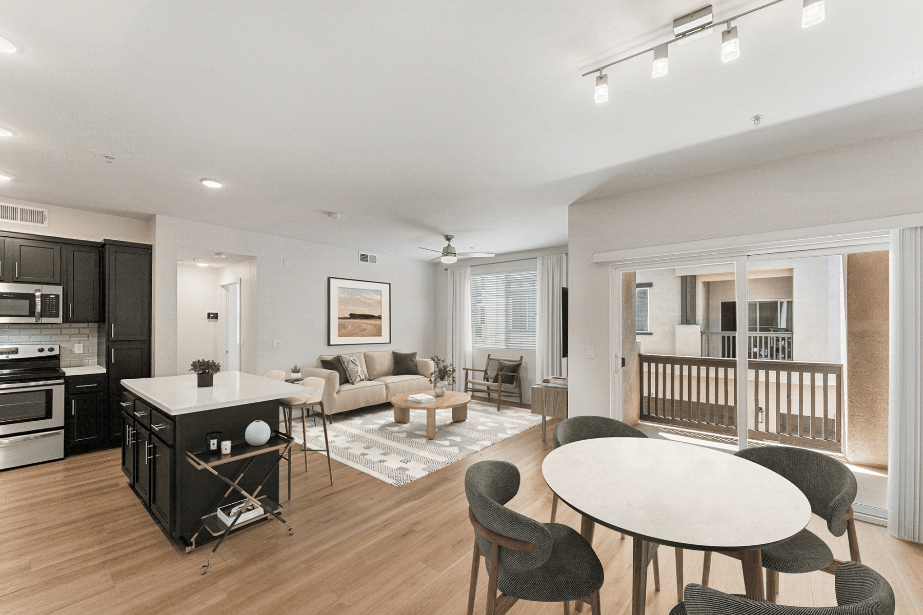 Open-concept kitchen, living room, and dining area featuring a kitchen island, beige sofa, round coffee table, and a dining table with four chairs overlooking a balcony.